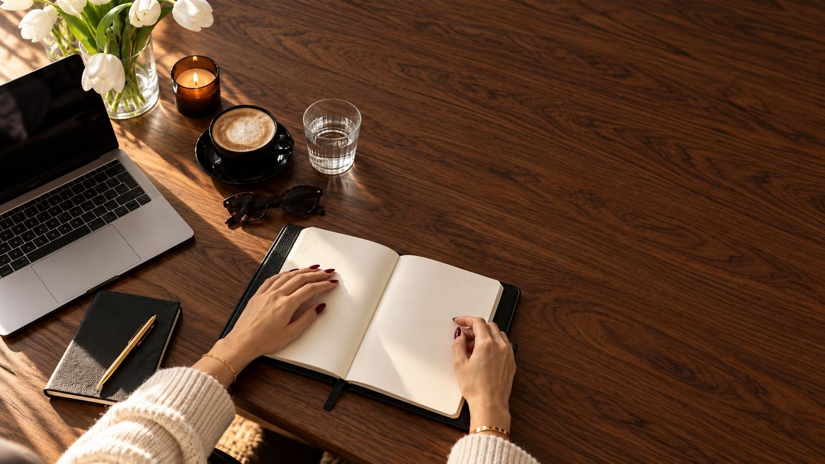 Hands with burgundy nails opening a journal beside a coffee on a dark wood table